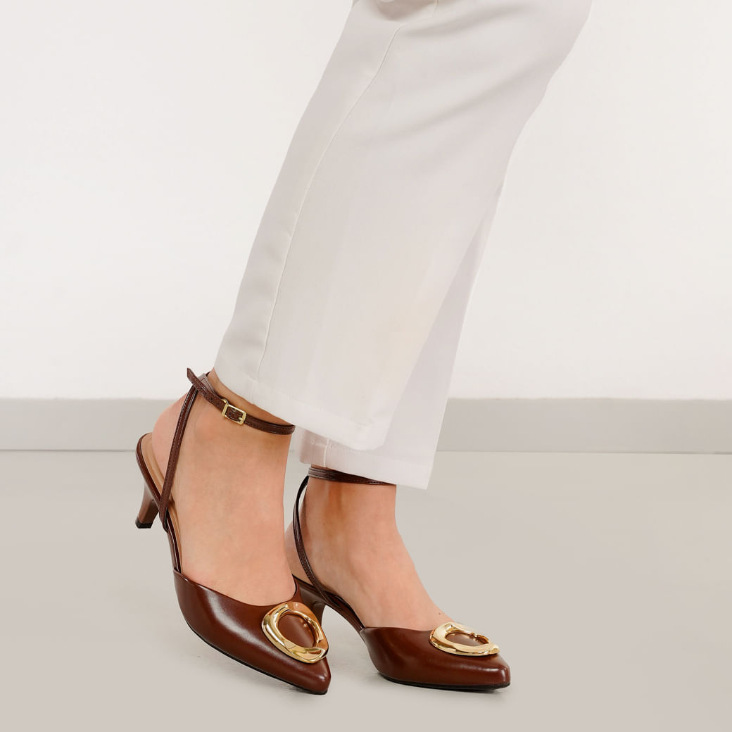 Scarpin feminino cor conhaque em posição com modelo sentado, bico fino, salto baixo, tira no tornozelo com fivela dourada e aplique metálico no cabedal.
